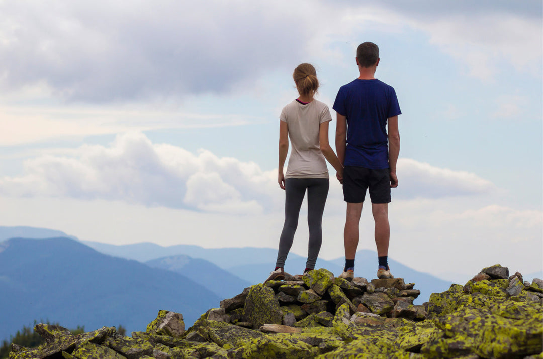 A couple stand on a mountaintop enjoying the view. They are dressed casually in summer clothing. 