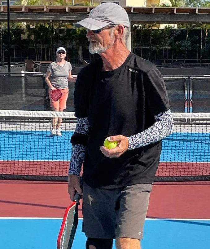Man wearing black and white sun  sleeves while holding a pickleball paddle and ball on a pickleball court with another player in the background.